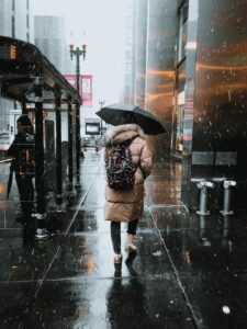 Woman in Rain by Mario Cuadros. Displays a woman in a city, walking along the sidewalk. She wears a long brown coat and a backpack. We see her walking away from us with a black umbrella over her head.