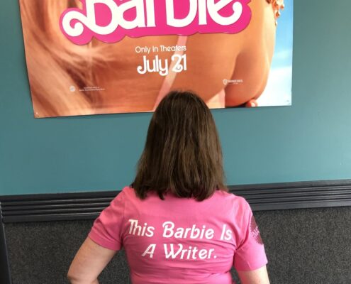 In front of the Barbie movie poster, with a t-shirt that says "This Barbie is a writer" on the back