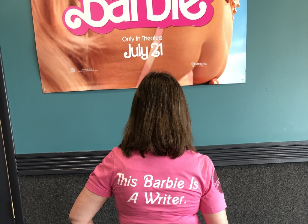 In front of the Barbie movie poster, with a t-shirt that says "This Barbie is a writer" on the back